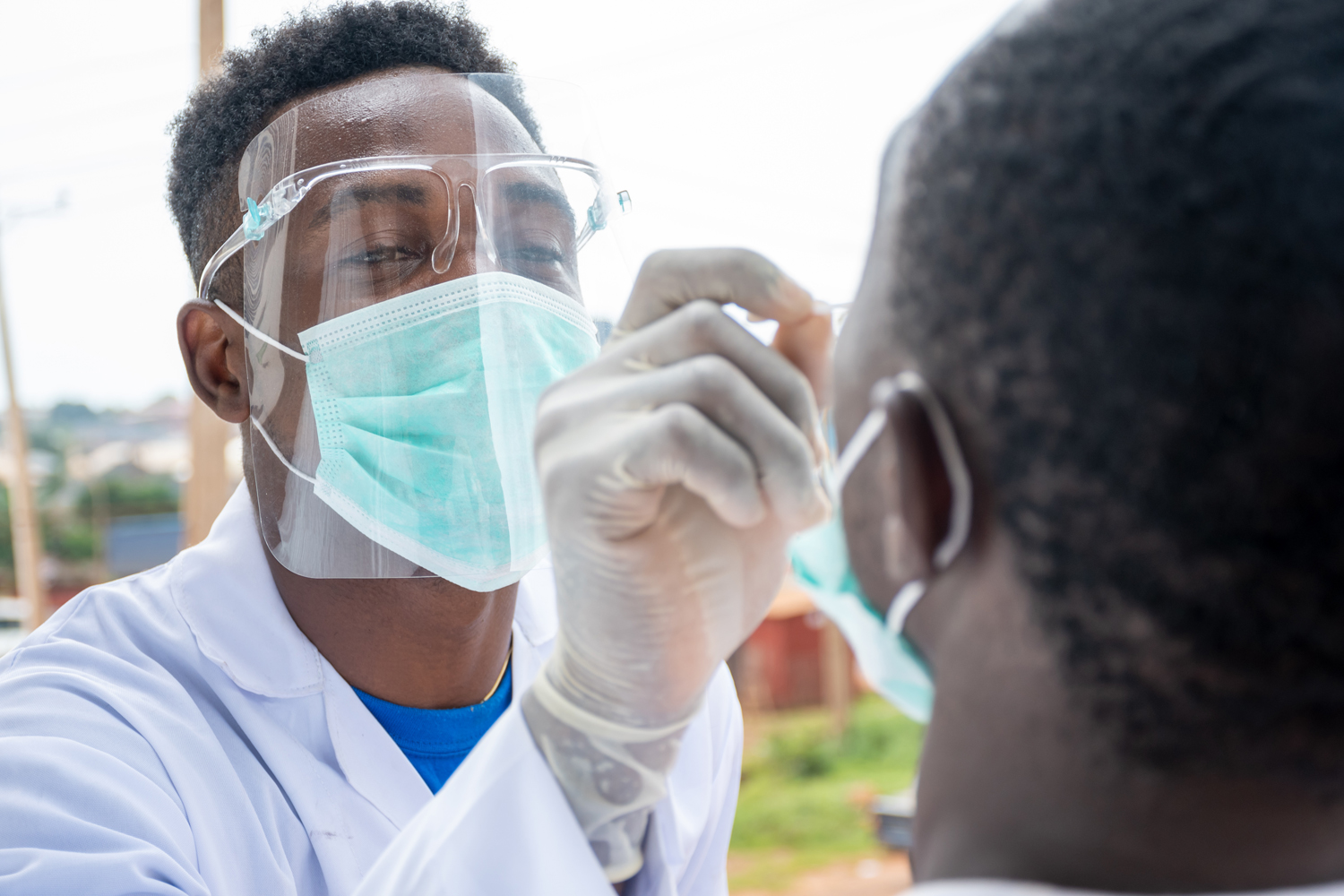 A young male doctor wears a surgical mask, shield, and gloves. He collects a nasal swab sample from a young male.