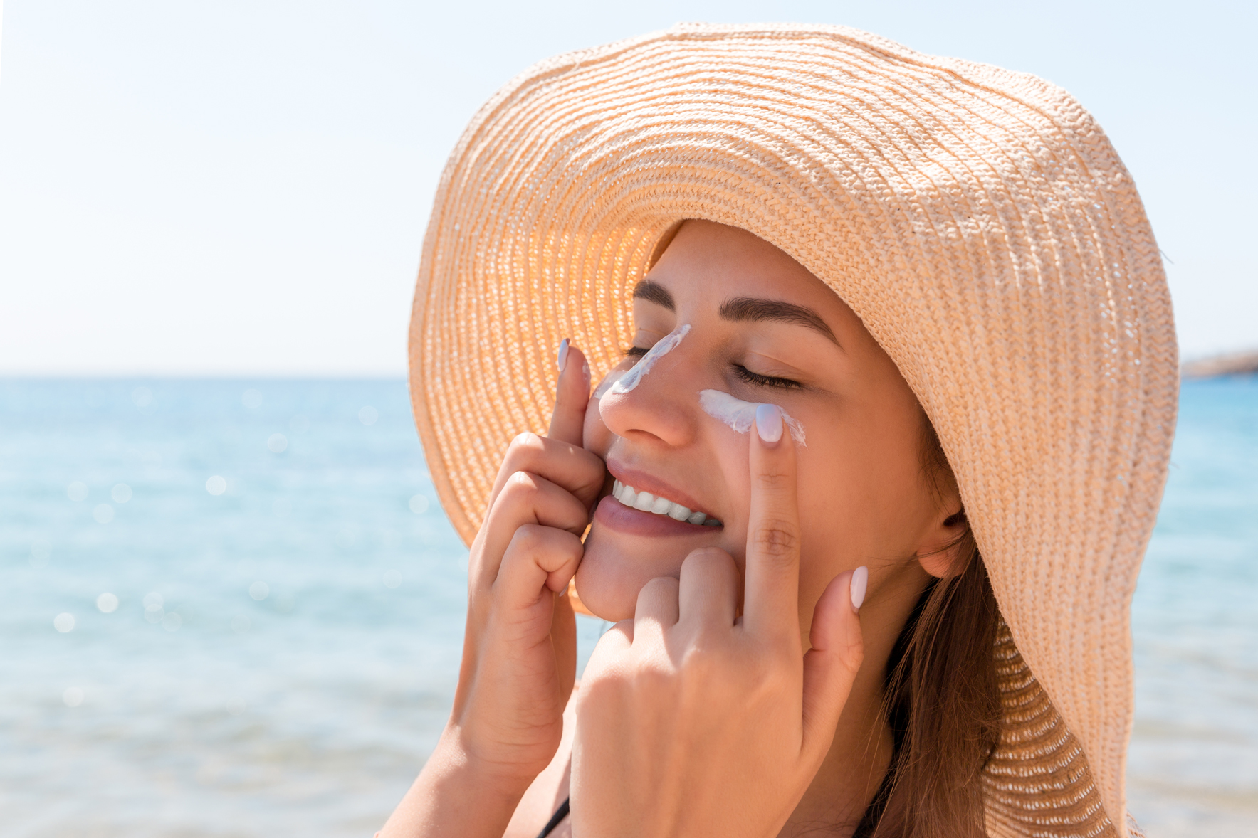 A smiling woman in a hat applies sunscreen to her face with a lake in the background.