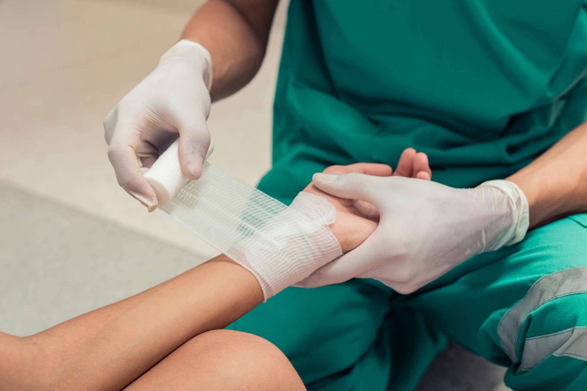 A closeup image of a medical professional in green scrubs and white gloves wrapping a wound on the wrist of a patient.