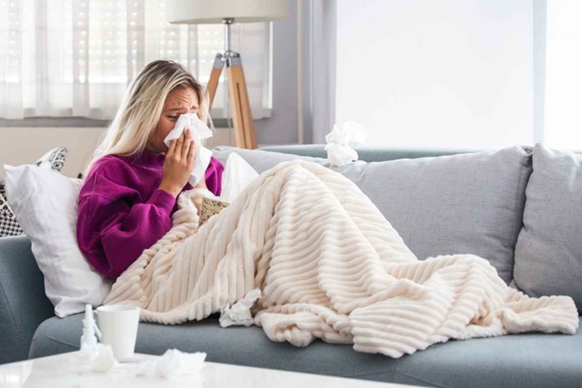 A blonde woman in a purple sweatshirt sits across her sofa, covered in a blanket, blowing her nose.