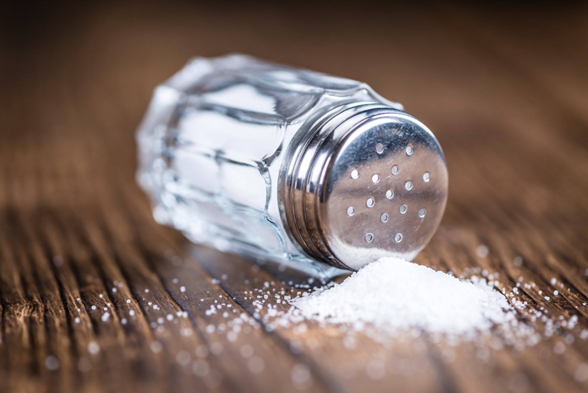 A closeup image of a tipped-over salt shaker against a wooden table.