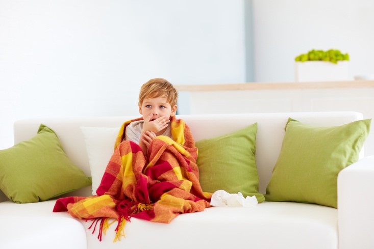 A sick young boy wrapped in a blanket sits on the sofa beside a pile of tissues while covering his cough.