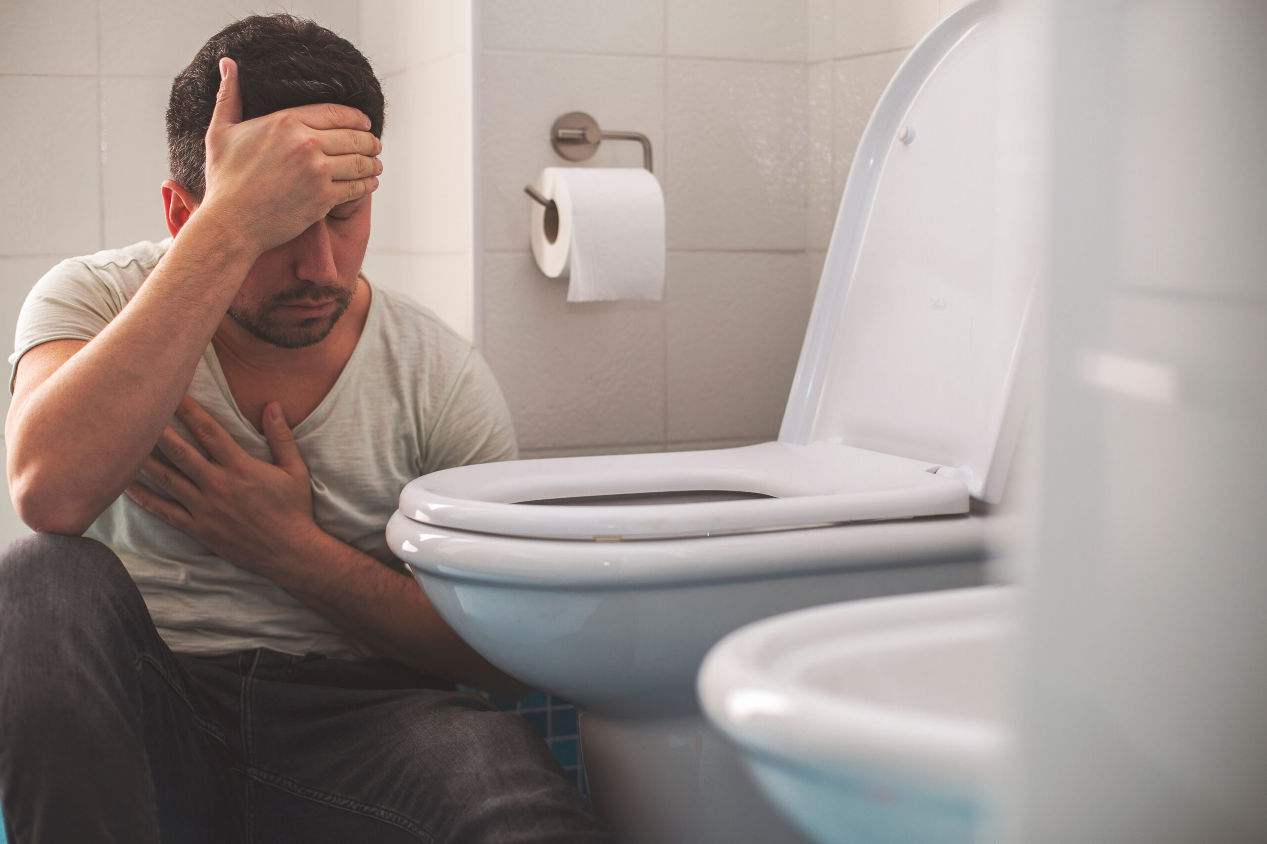 A man sits on the floor in front of a toilet holding his head and chest in pain.