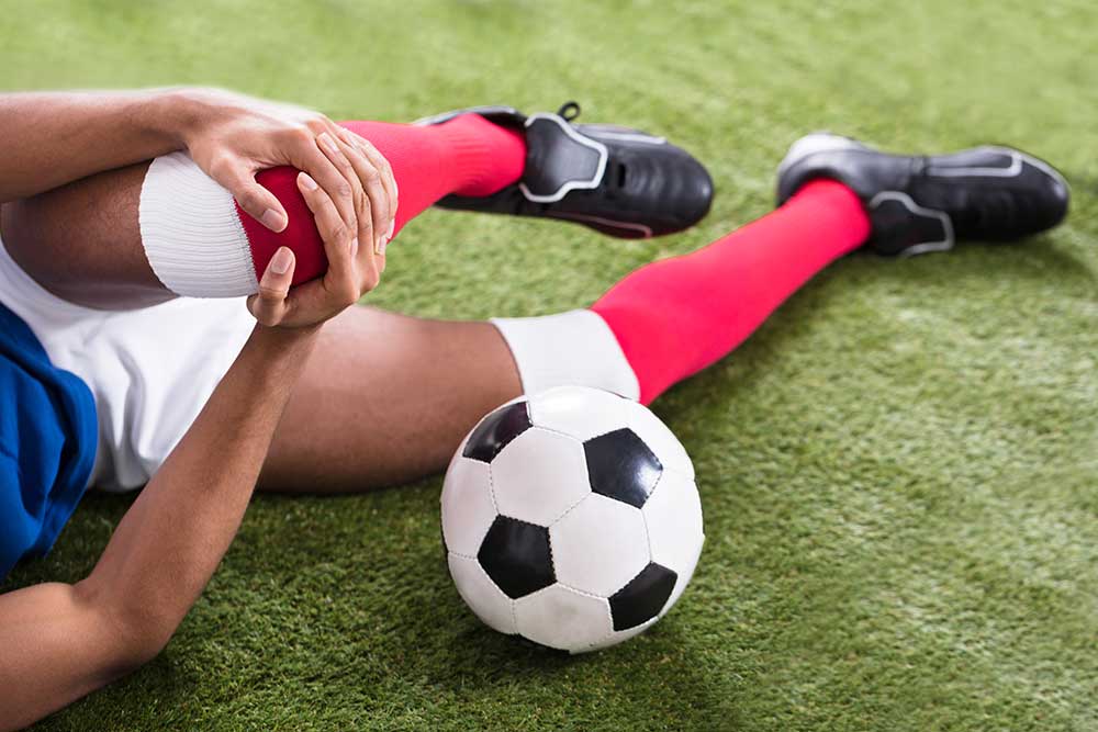 A closeup image of a male soccer player on the turf gripping his knee with his hands near a soccer ball.