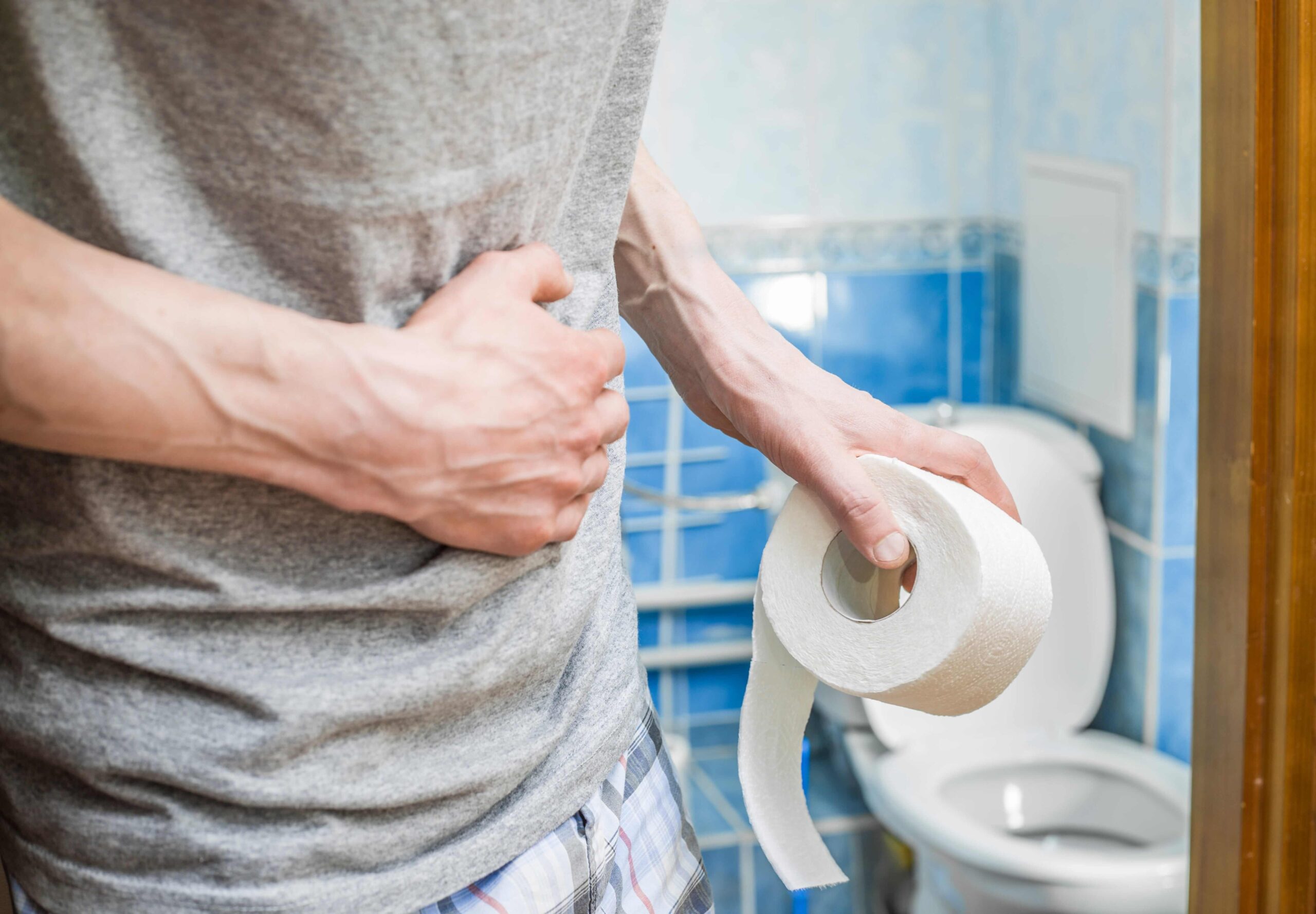 A closeup of a man in the bathroom holding a roll of toilet paper in one hand and gripping his stomach in pain with the other.